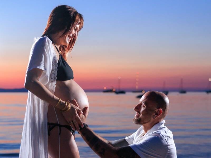 Un homme à genoux devant sa femme enceinte pose ses mains sur son ventre rond, avec un coucher de soleil sur la mer en toile de fond.