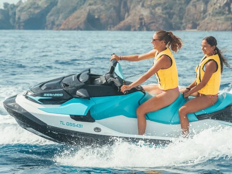 Une femme et une jeune fille vêtues de gilets de sauvetage jaunes sont sur un Jet Ski bleu turquoise, blanc et gris en marche.