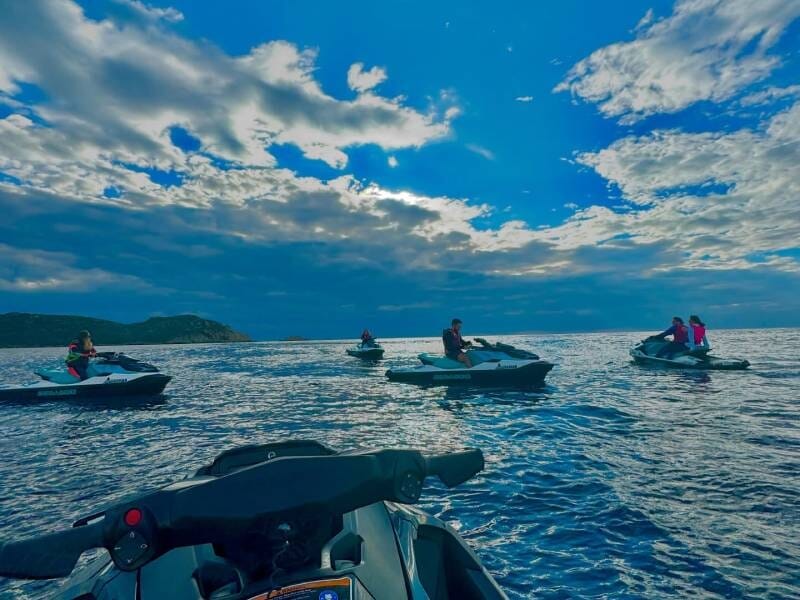 Vue sur la mer et 5 jet skis avec des passagers, le ciel est bleu avec des nuages blancs.