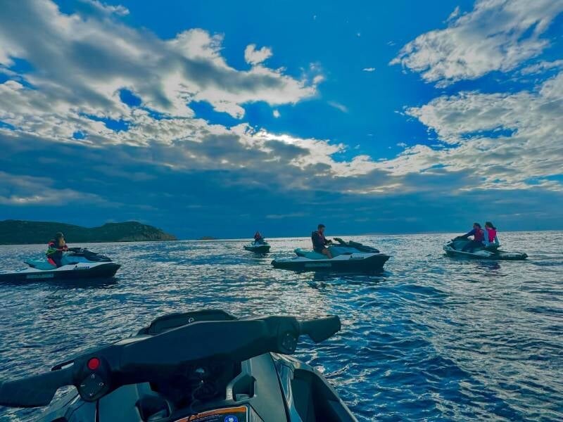 Vue sur la mer et 5 jet skis avec des passagers, le ciel est bleu avec des nuages blancs.