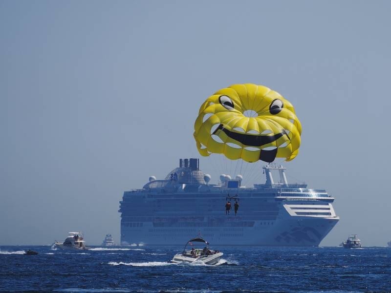 Un bateau traîne un parachute ascensionnel jaune avec des yeux et un sourire dans le ciel. En fond se trouve un gigantesque paquebot.