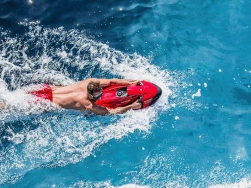 Un homme accroché à un propulseur sous-marin rouge, dans une eau turquoise.
