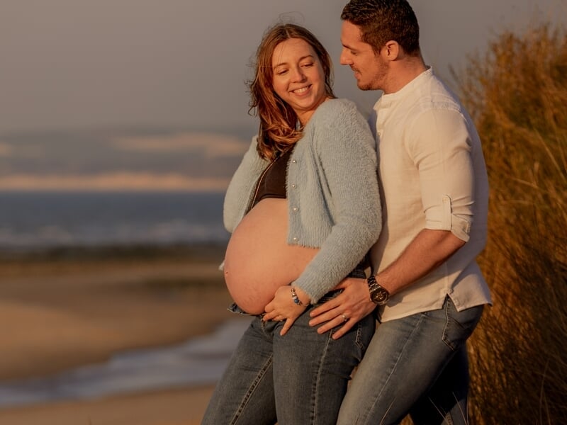 Couple souriant sur la plage au coucher du soleil, femme enceinte en pull bleu, lors d'un shooting avec une photographe grossesse sur la Côte d'Opale.