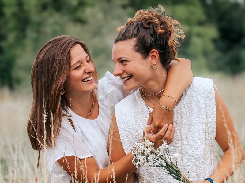 Deux jeunes femmes souriantes dans un champ, l'une tenant un bouquet de fleurs blanches, ambiance naturelle et joyeuse.