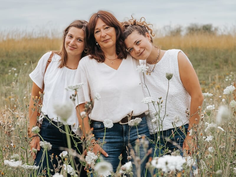 Trois femmes souriantes posent ensemble dans un champ de fleurs sauvages sous un ciel nuageux.