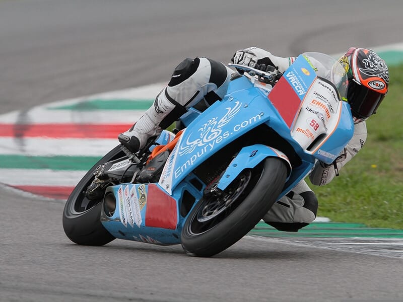 Pilote en combinaison blanche et noire prenant un virage serré sur une moto de course bleue Empuryes.