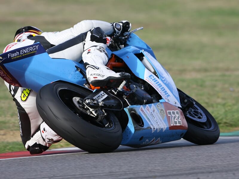 Motard penché dans un virage sur circuit, au guidon d'une moto bleue Empuryes sponsorisée par Flash Energy.