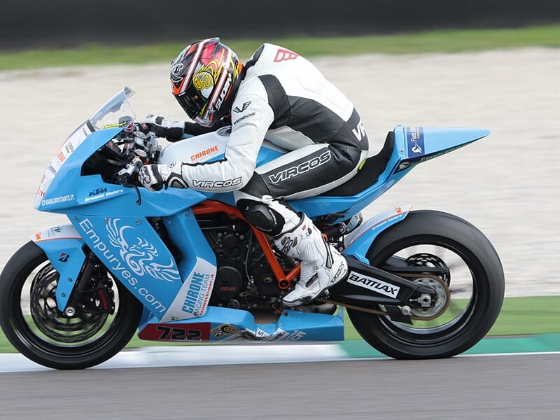 Pilote concentré, en combinaison blanche et noire, sur une moto bleue Empuryes numéro 722 en pleine accélération.