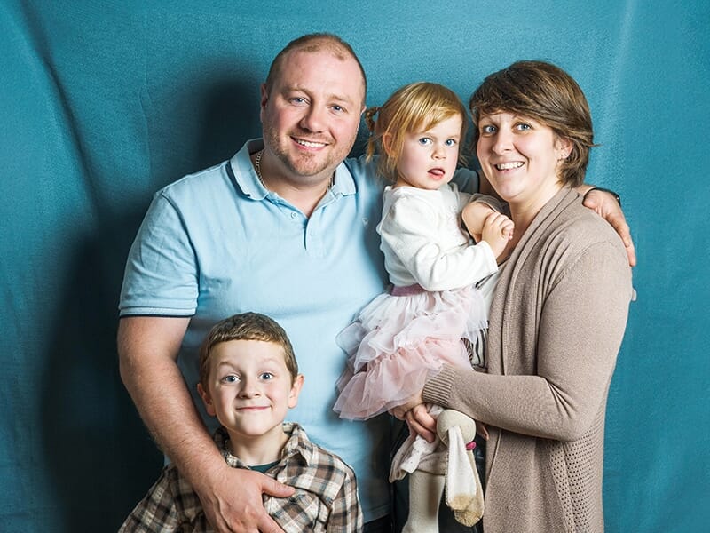 Portrait de famille avec deux parents souriants et leurs deux jeunes enfants devant un fond bleu.