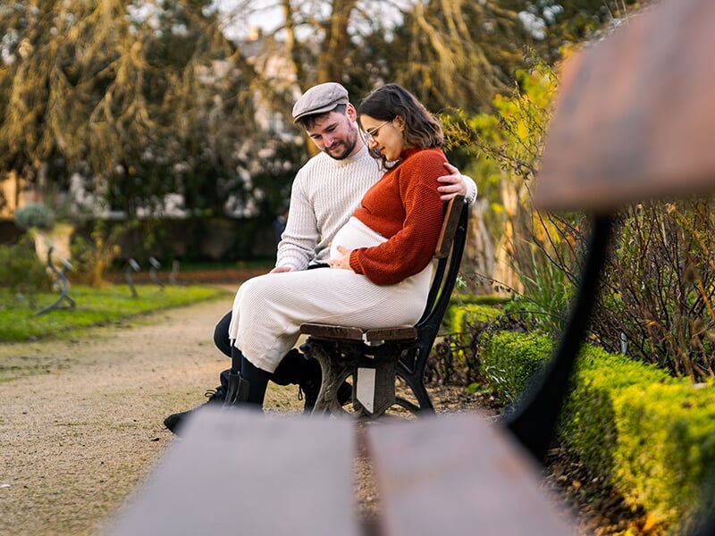 Couple assis sur un banc dans un parc, regardant affectueusement le ventre de la future maman.