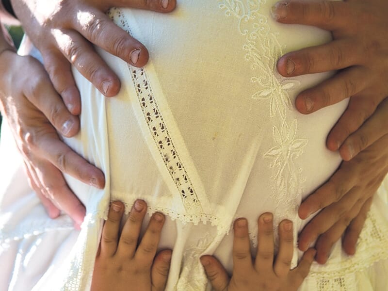 Plusieurs mains d’adultes et d’enfants posées sur le ventre arrondi d’une femme enceinte vêtue de blanc.