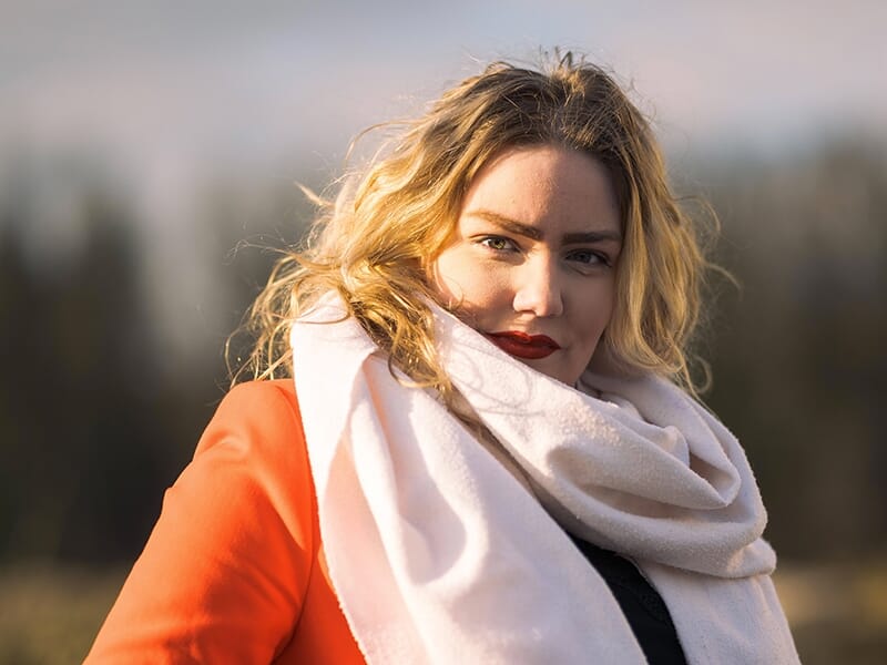 Femme blonde portant un manteau orange et une grande écharpe blanche, éclairée par la lumière dorée du soleil.