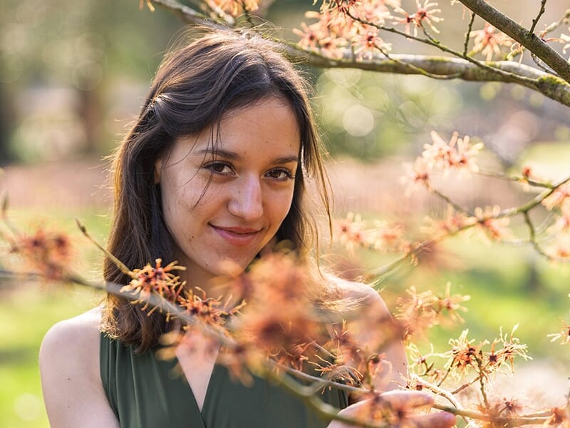 Jeune femme souriante au milieu de branches fleuries dans un jardin baigné de lumière naturelle.