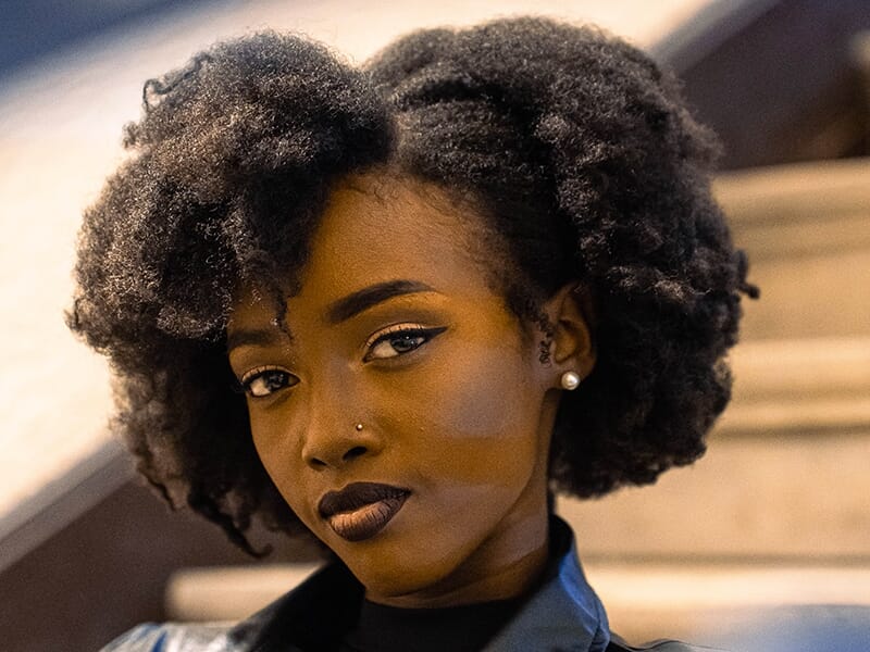 Femme au regard intense avec coiffure afro naturelle, maquillage soigné et piercing au nez.