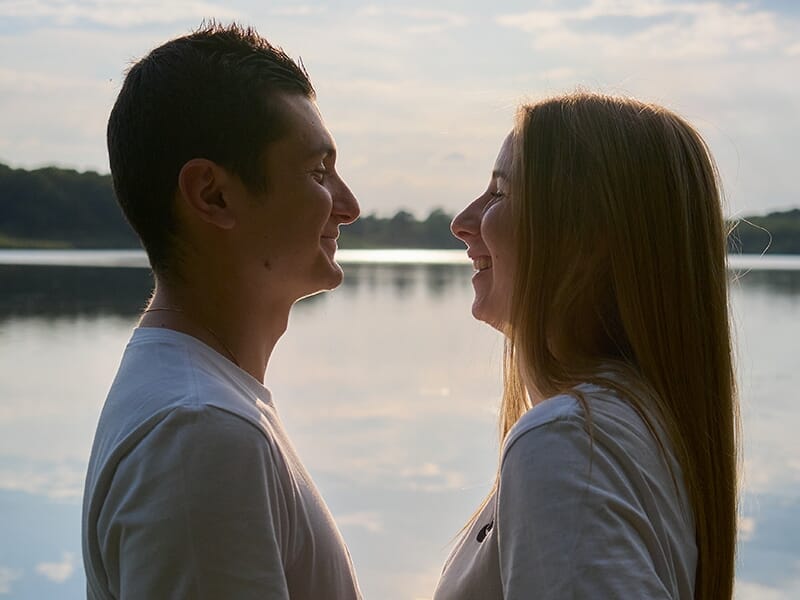 Couple face à face au bord d’un lac au coucher du soleil, souriants et complices.