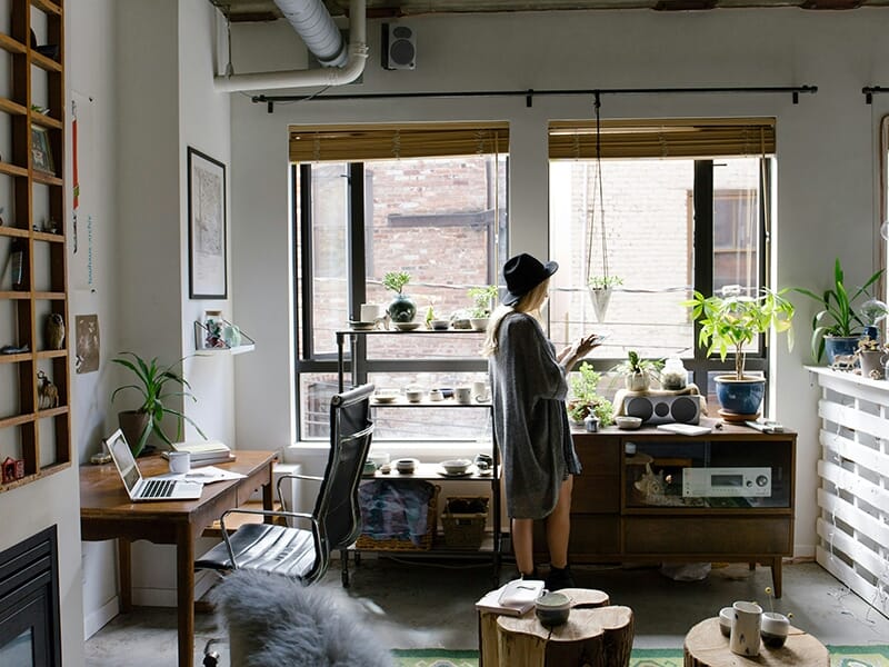 Femme portant un chapeau, debout devant une fenêtre, s’occupant de plantes dans un intérieur cosy.