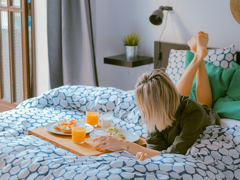 Femme allongée sur un lit avec un plateau de petit-déjeuner, dans une chambre lumineuse et décorée.