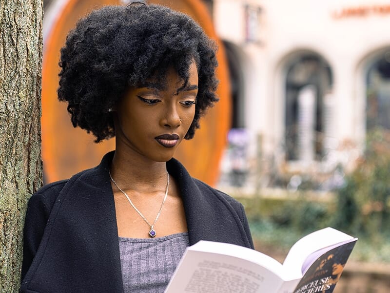 Femme lisant un livre adossée à un arbre, concentrée sur sa lecture.