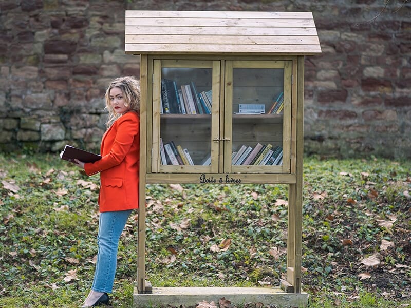 Femme en veste rouge tenant un livre à côté d'une boîte à livres en bois dans un parc.