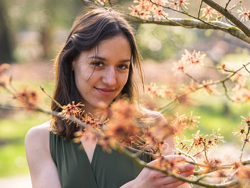 Femme souriante en robe verte posant derrière des branches fleuries dans un parc.