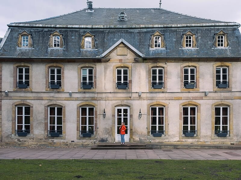 Femme debout devant une grande bâtisse ancienne avec de nombreuses fenêtres.