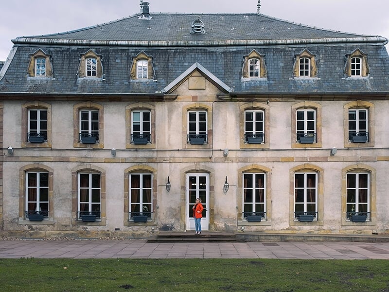 Femme debout devant une grande bâtisse ancienne avec de nombreuses fenêtres.