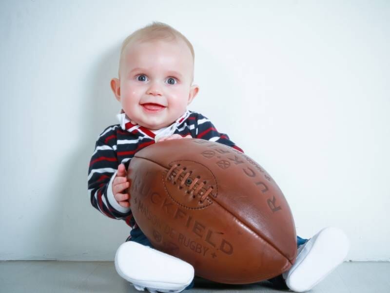 Un bébé souriant prend la pause avec un ballon de rugby devant lui.