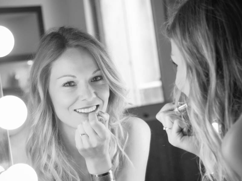 Photo en noir et blanc d'une jeune femme blonde qui met du rouge à lèvre devant un miroir et dont le reflet sourit à l'objectif.