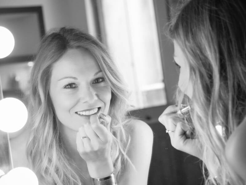 Photo en noir et blanc d'une jeune femme blonde qui met du rouge à lèvre devant un miroir et dont le reflet sourit à l'objectif.