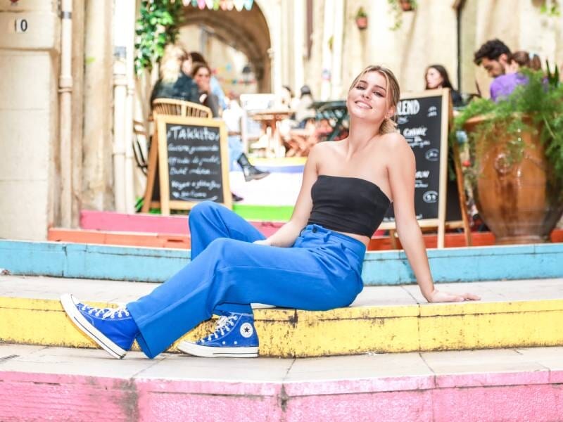 Une jeune femme avec un haut noir, un pantalon et des baskets bleus prend la pose en pleine rue, assise sur des escaliers multicolors.