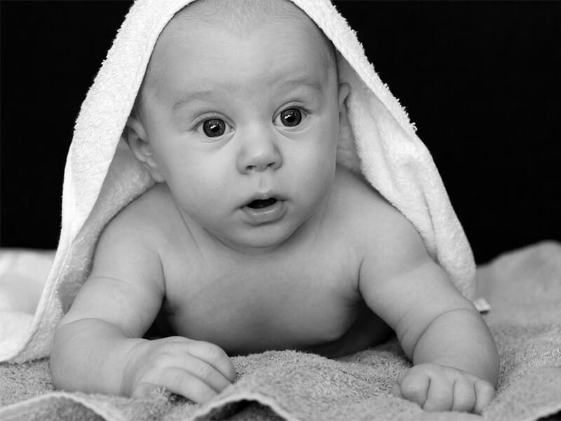 Bébé allongé sur une serviette, regard curieux, partiellement couvert d’une serviette blanche sur la tête.