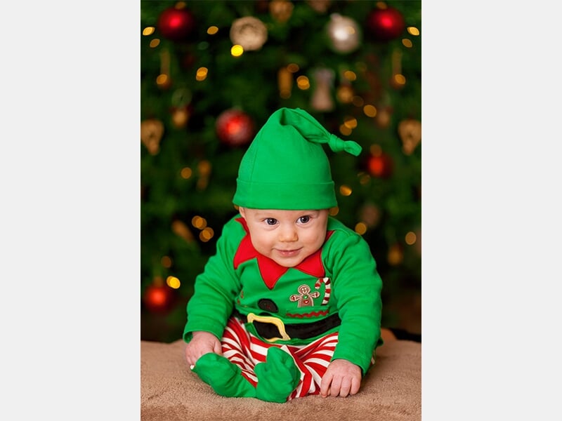 Bébé déguisé en lutin de Noël avec bonnet vert, assis devant un sapin décoré de boules rouges et dorées.