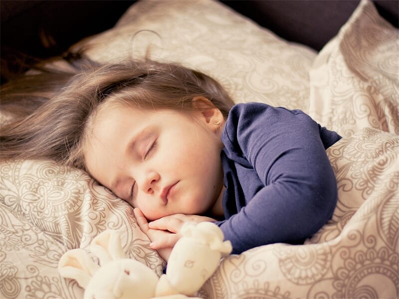 Jeune enfant endormi sur un oreiller à motifs, peluche dans les bras, vêtue d’un haut violet à manches longues.