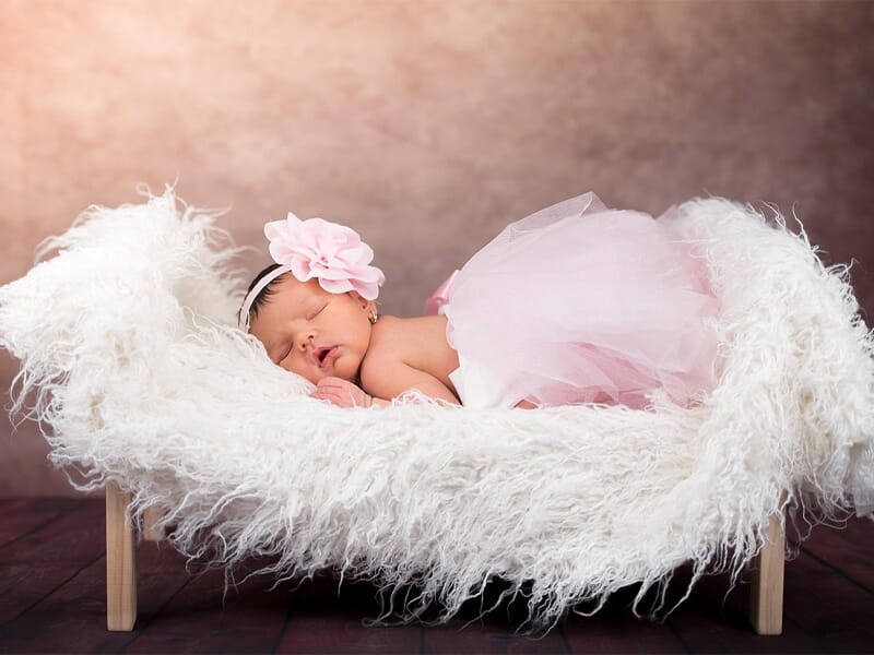Nouveau-né endormi dans un petit lit en fourrure blanche, robe rose en tulle et large fleur rose sur le bandeau.