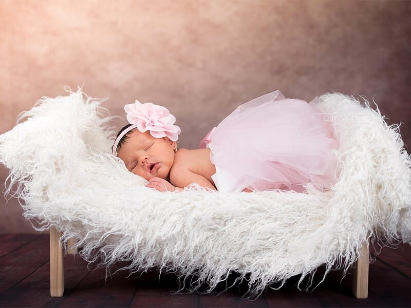 Nouveau-né endormi dans un petit lit en fourrure blanche, robe rose en tulle et large fleur rose sur le bandeau.