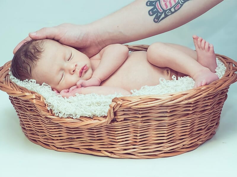 Bébé dormant dans un panier en osier, main adulte tatouée posée sur sa tête, couverture crème en dessous.