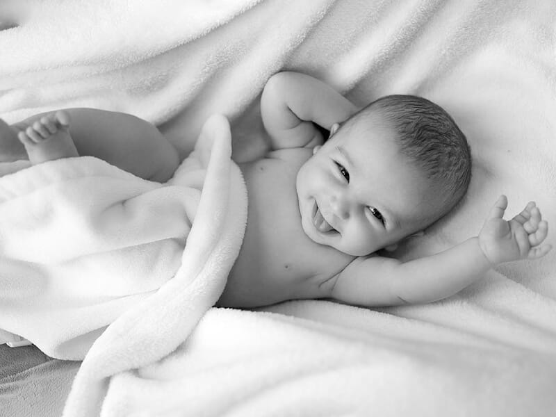 Bébé souriant allongé sur une couverture blanche, bras levés, langue tirée, regard tourné vers l’objectif.
