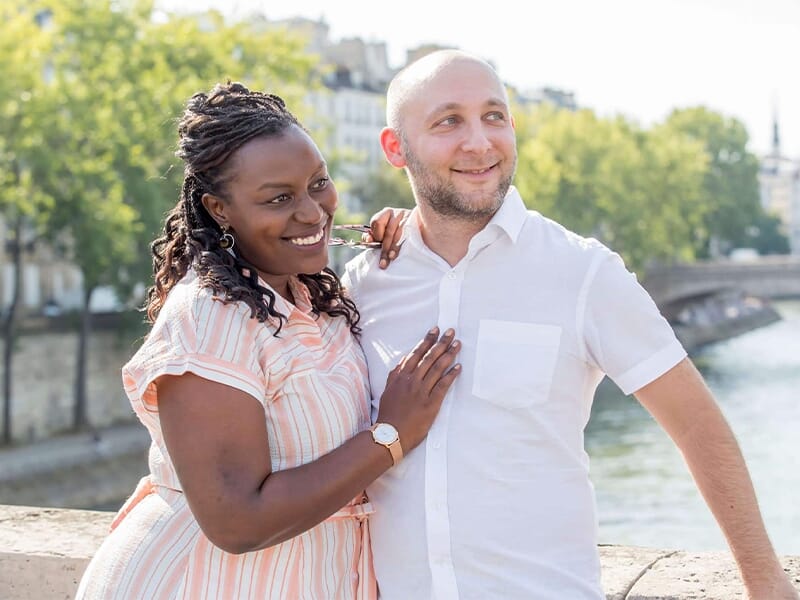 Couple souriant enlacé près d’un fleuve, lumière naturelle, arrière-plan urbain et verdoyant.