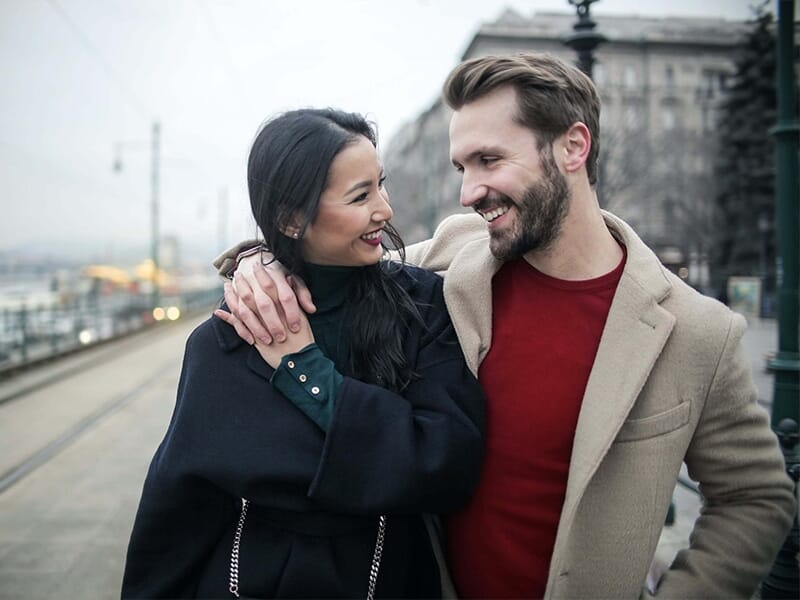 Couple souriant marchant bras dessus bras dessous en manteaux, regard complice dans une rue urbaine.