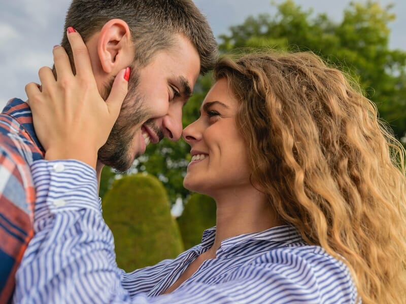 Couple en gros plan, front contre front, regard tendre, arbres en arrière-plan par temps clair.