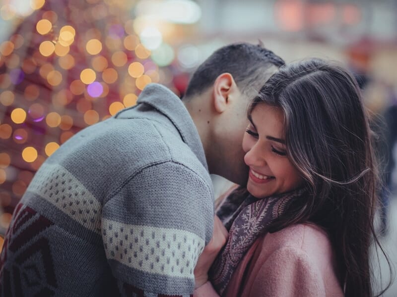 Homme chuchotant à l’oreille d’une femme souriante, décor de Noël flou en arrière-plan.