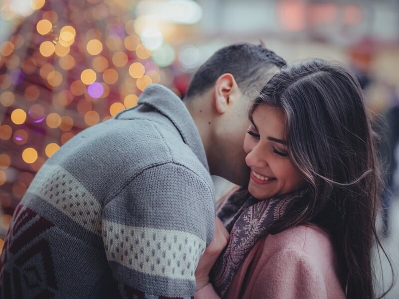 Homme chuchotant à l’oreille d’une femme souriante, décor de Noël flou en arrière-plan.