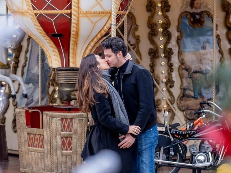 Couple s’embrassant devant un manège décoratif, ambiance festive avec montgolfière peinte et lumières.