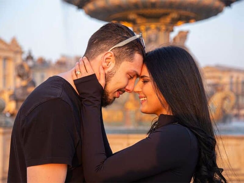 Couple touchant leurs fronts en souriant devant une fontaine décorée, lumière dorée du coucher de soleil.