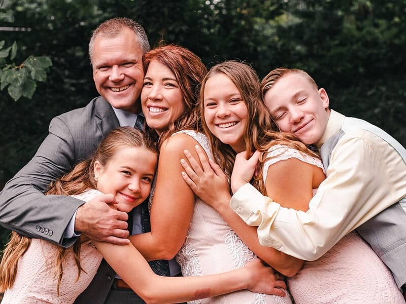 Famille de cinq personnes se serrant dans les bras avec de larges sourires, fond boisé et tenues habillées.