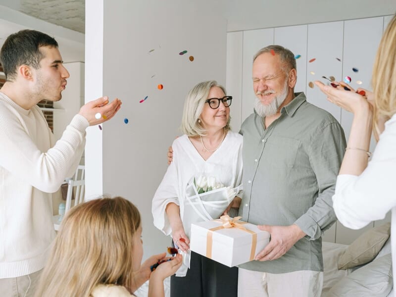Couple de personnes âgées entouré de membres de la famille lançant des confettis, avec bouquet et cadeau en main.
