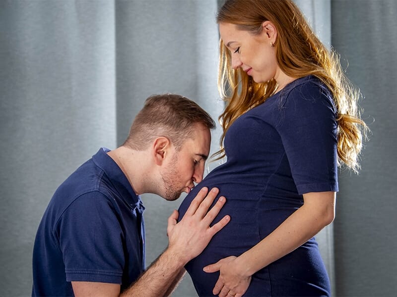 Homme embrassant tendrement le ventre rond de sa compagne enceinte, tous deux en vêtements bleu marine.