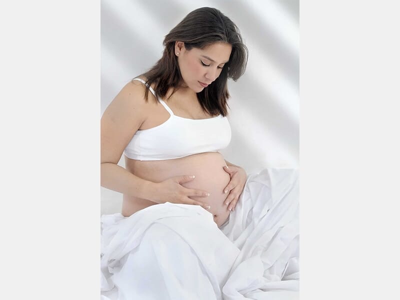Femme enceinte assise sur un lit, habillée d’un haut blanc, regard baissé sur son ventre nu.