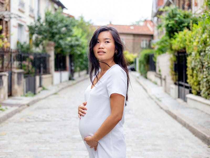 Femme enceinte en robe blanche tenant son ventre dans une rue pavée bordée de maisons et de verdure.