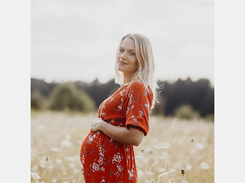 Femme enceinte debout dans un champ fleuri, portant une robe rouge à motifs floraux, regard tourné vers l’objectif.